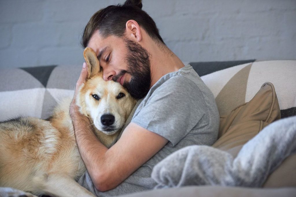 Un homme blanc aux cheveux noir qui fait un câlin a son chien beige qui pleur. Ils sont dans le sofa