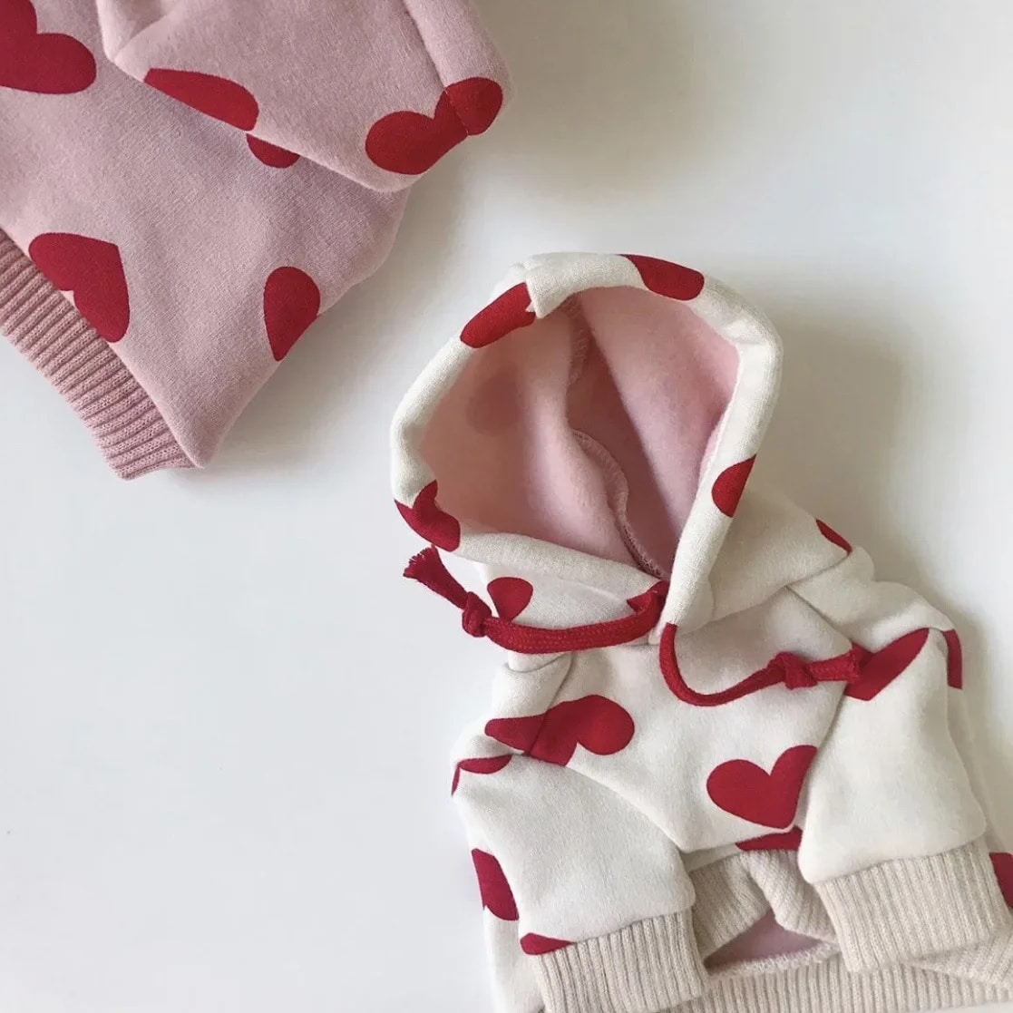 Deux pulls sweat à capuche pour chien dont un blanc et un rose avec des motifs de coeurs rouges.