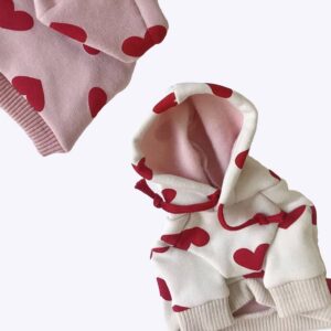 Deux pulls sweat à capuche pour chien dont un blanc et un rose avec des motifs de coeurs rouges.
